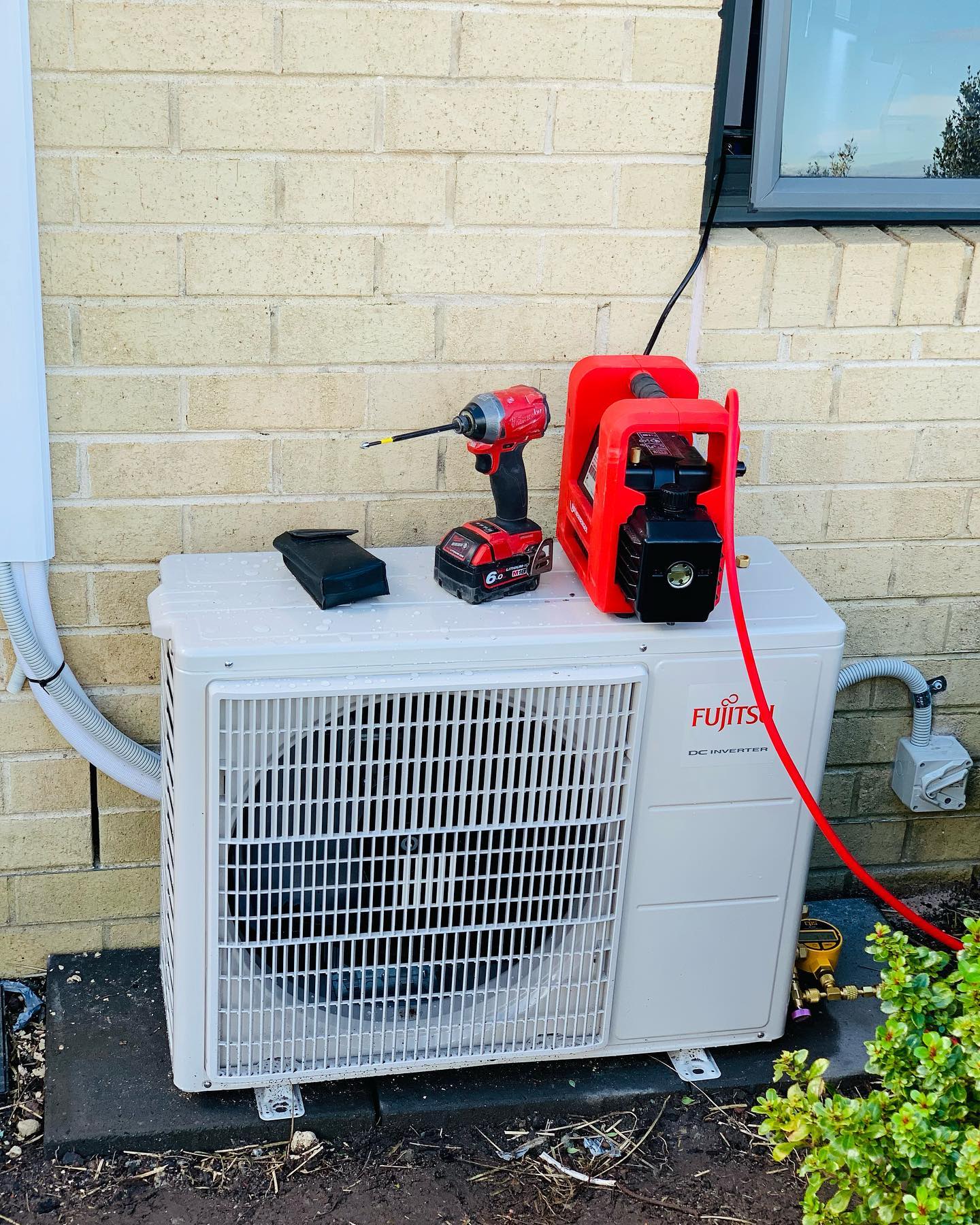 A Fujitsu DC Inverter outdoor unit newly mounted on a brick wall, with a vacuum pump and impact driver sitting on top mid-install