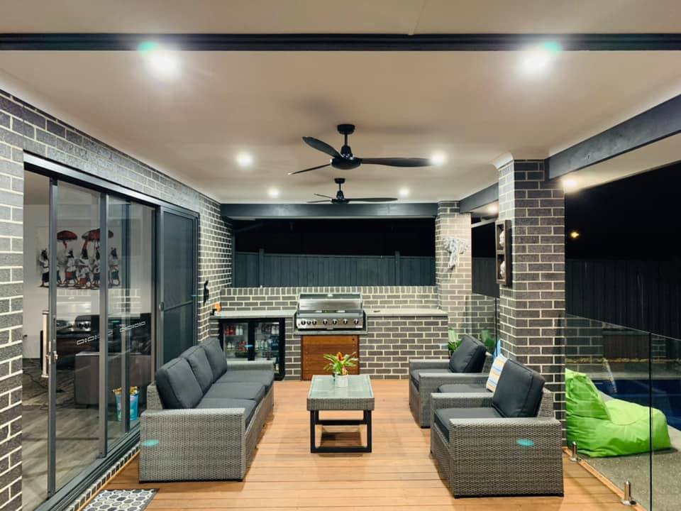 An alfresco area at night lit by warm downlights, with two black ceiling fans above an outdoor kitchen and lounge