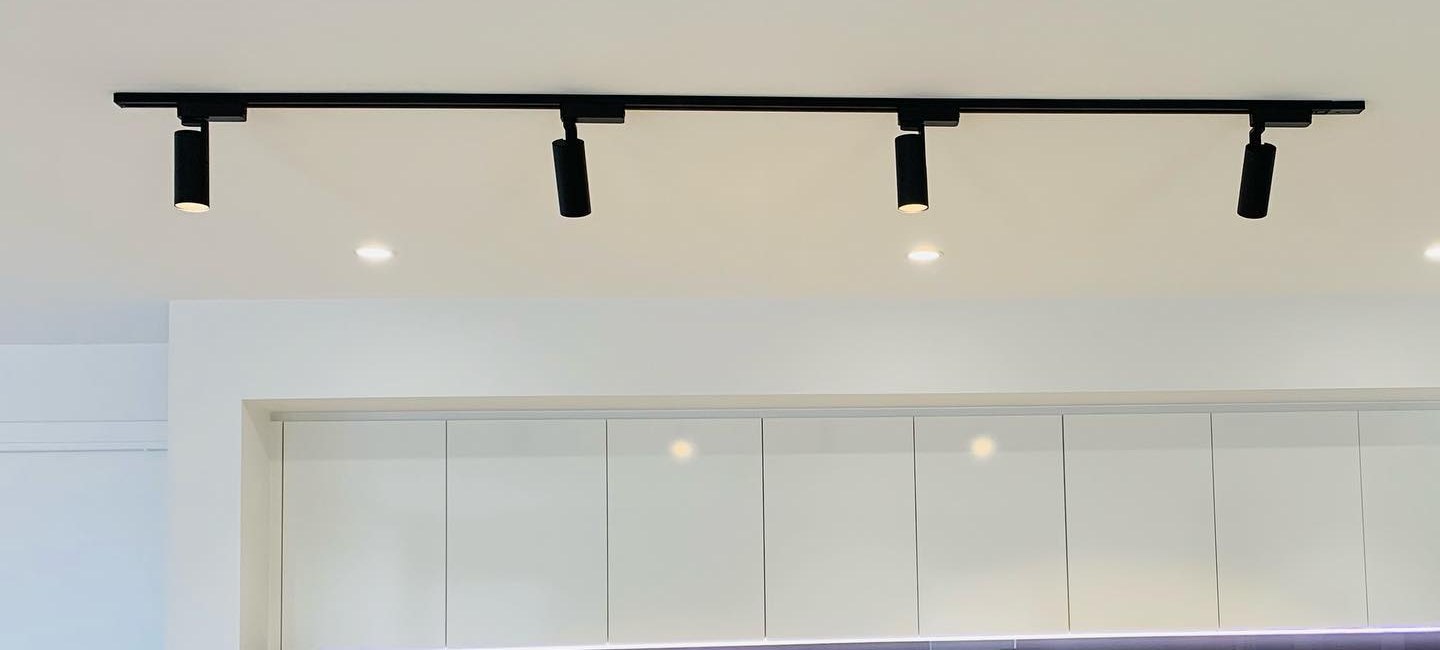 A long black ceiling track with four spotlights above white kitchen cabinetry, with downlights between